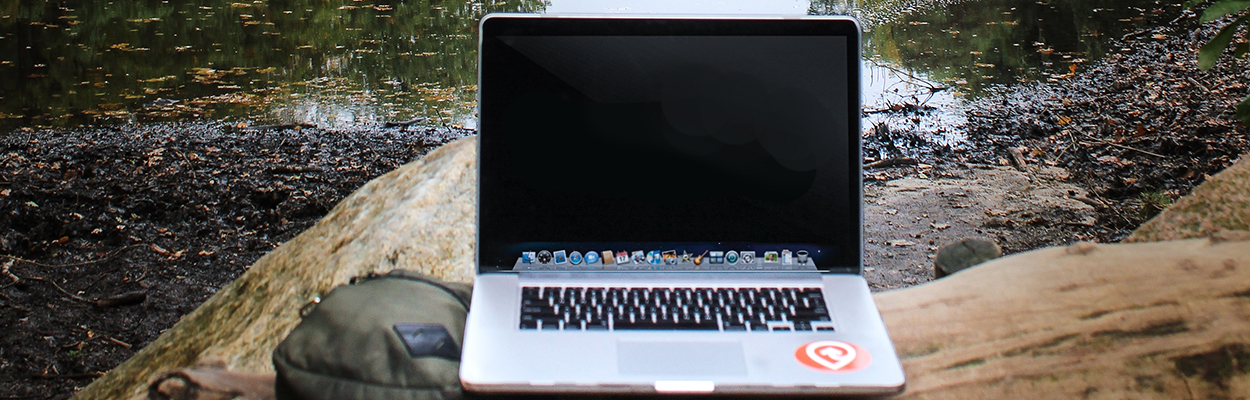 An open Mac laptop on a rock next to a backpack with a river in the background.