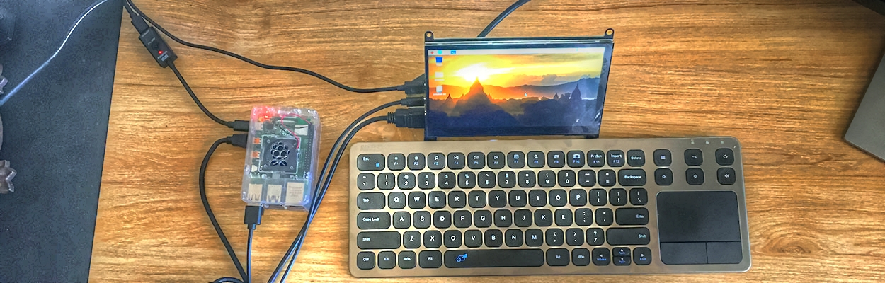 a raspberry pi in a clear case beside a silver keyboard with black keys and a 7-inch touchscreen monitor, as viewed from above, on a wooden desk