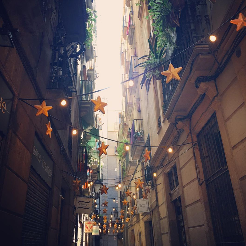 a narrow street in Barcelona with plants on balconies and string lights with stars going across